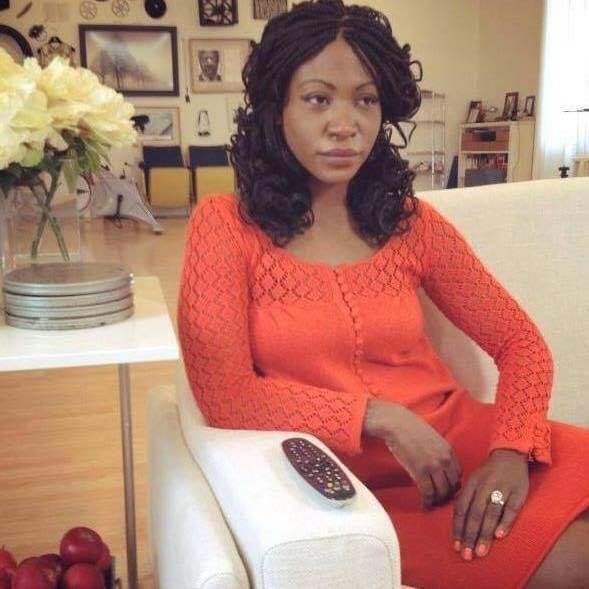 Africa Yoon dressed in an orange dress sitting on a white couch next to a white side table with white flowers and stack of vintage tin 8mm film reel canisters. In the background, is a wall of black and white photos.