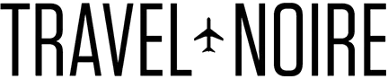 TRAVEL NOIRE logo is tall thin black sans-serif font and between the two words is a silhouette icon of a plane or airliner flying north.